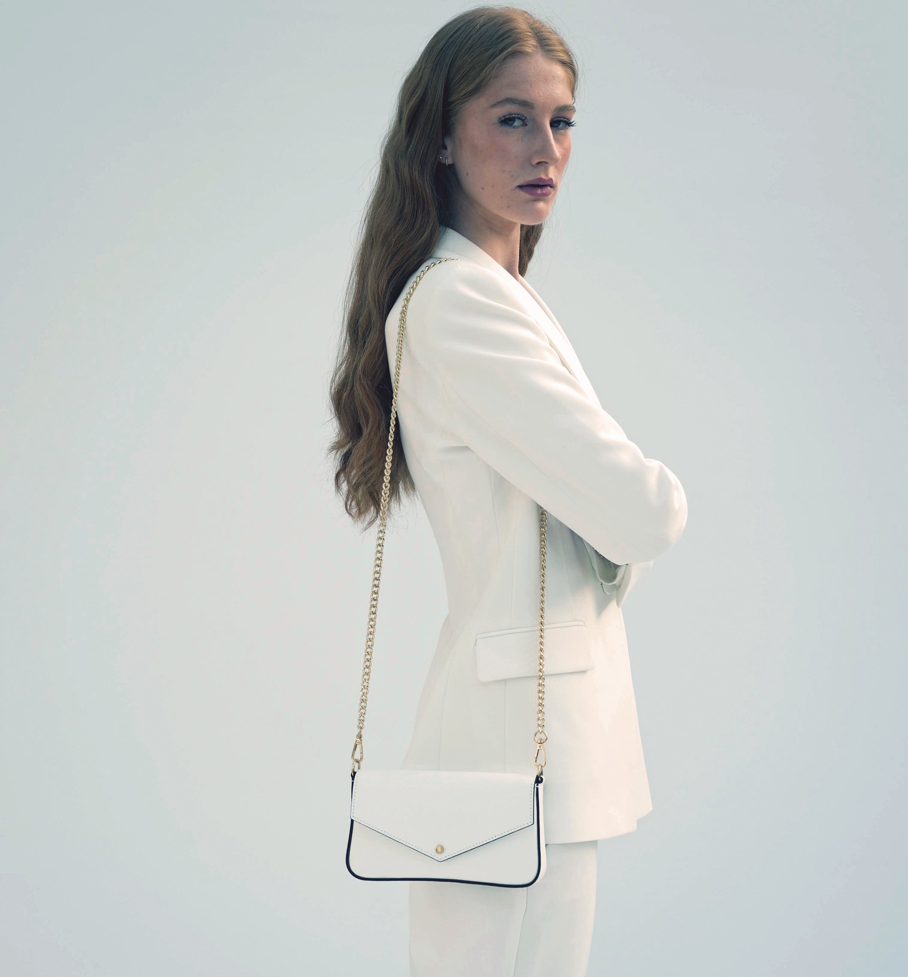 Woman in a white suit holding a white handbag with a gold chain strap against a light grey background